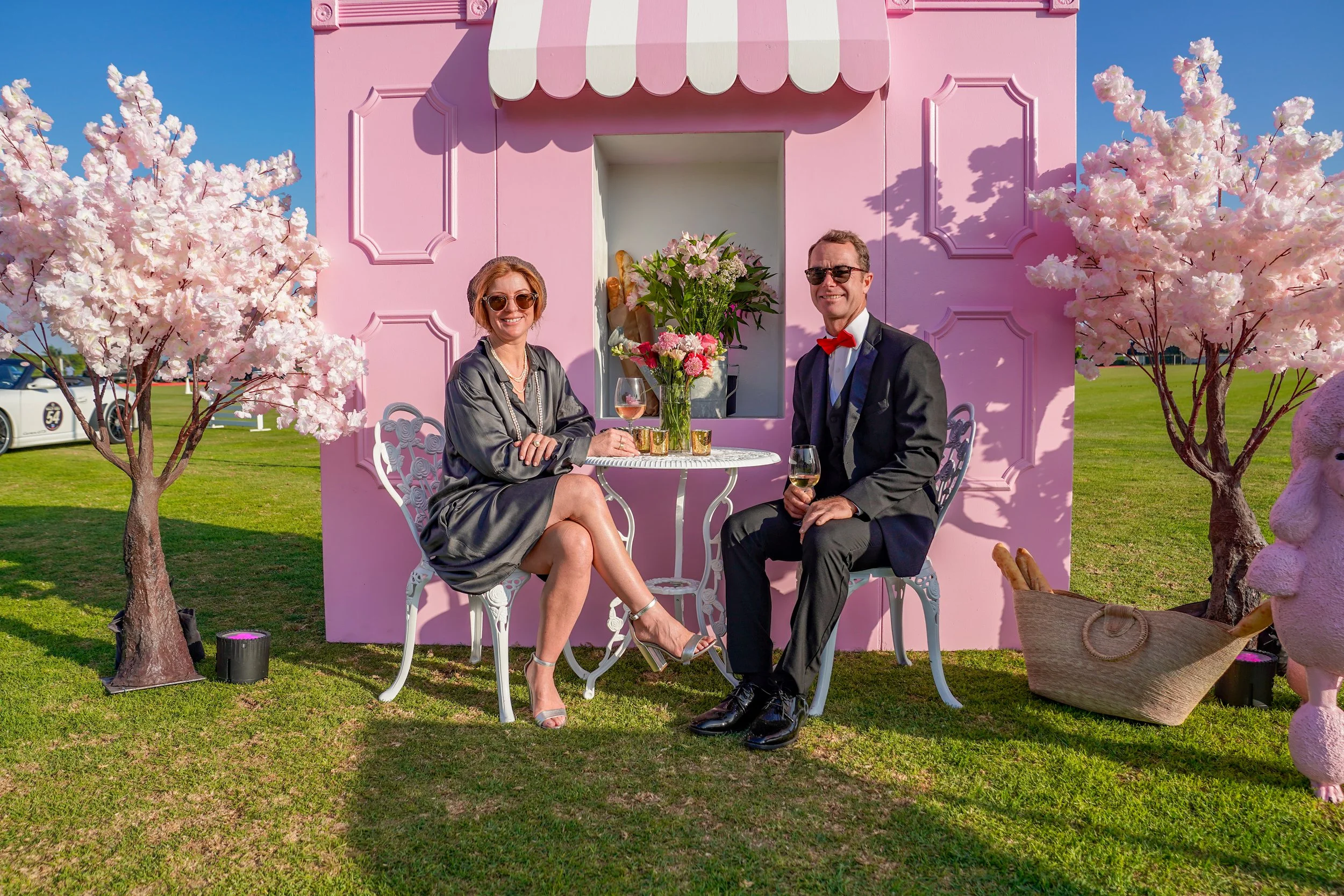 A woman and a man sitting at a small round table with pink flowers in a vase, two glasses of wine, in front of a pink backdrop with decorative panels and an awning. They are outdoors on a grassy area, each smiling and wearing sunglasses. The woman is