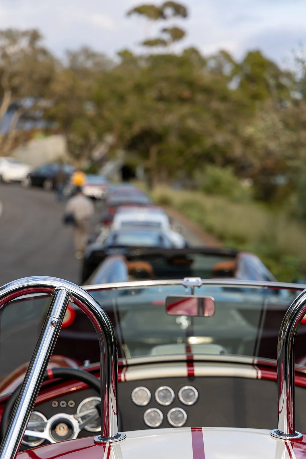View from the driver's seat of a red and white racing car looking through the windshield at a line of parked classic cars and people walking along a road.