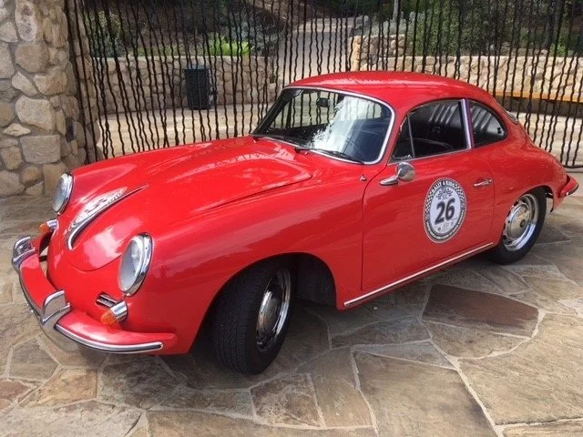 Red vintage Porsche 911 with racing number 26 decal, parked on stone pavement near a stone wall and black fence.