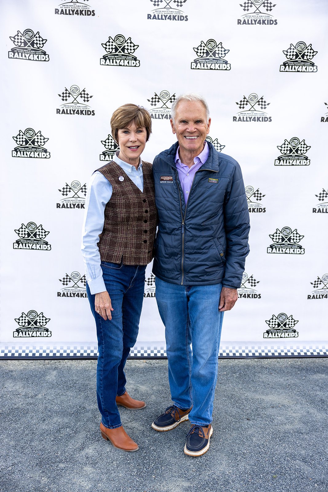 Two people standing in front of a backdrop with the "Rally4Kids" logo. The woman is wearing a brown plaid vest, blue shirt, jeans, and tan boots. The man is wearing a purple shirt, navy jacket, jeans, and boat shoes. Both are smiling.