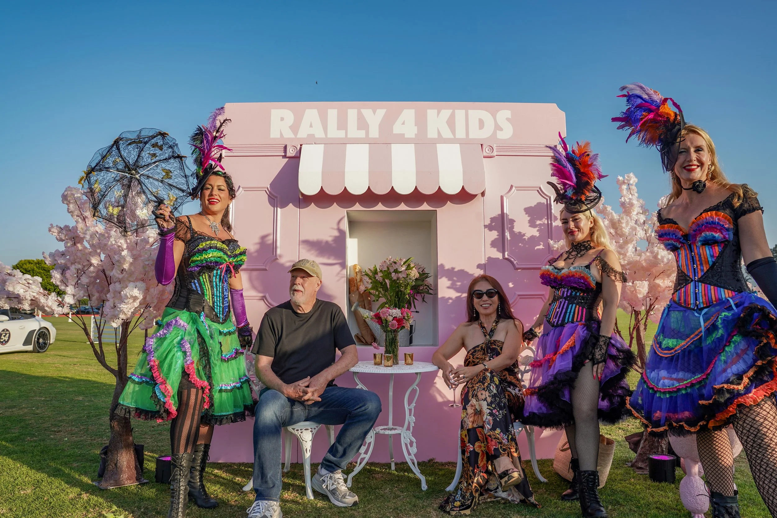People dressed in colorful costumes at a pink booth labeled "Rally 4 Kids" with cherry blossom trees and vintage cars in the background.