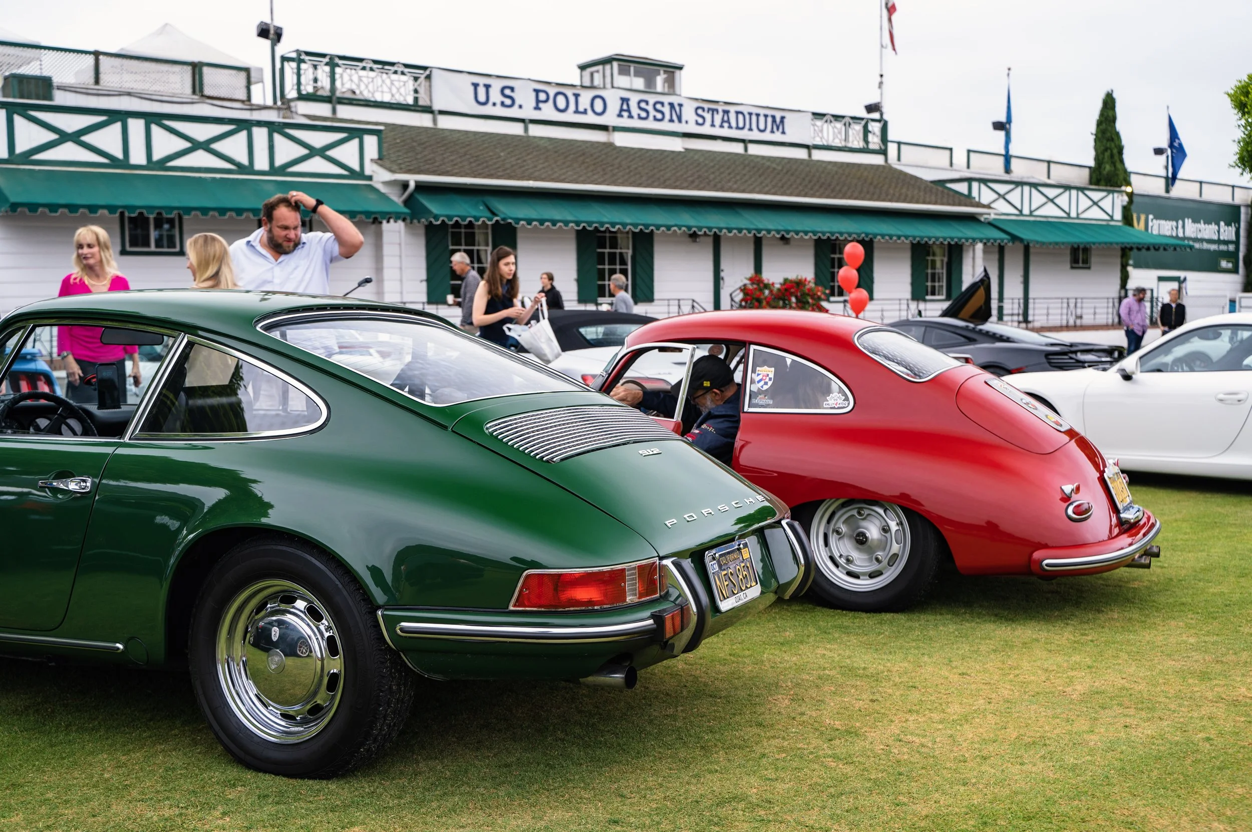 A classic car show in front of the U.S. Polo Assn. Stadium, featuring green and red vintage Porsche cars on a grassy area, with people walking and looking at the cars.