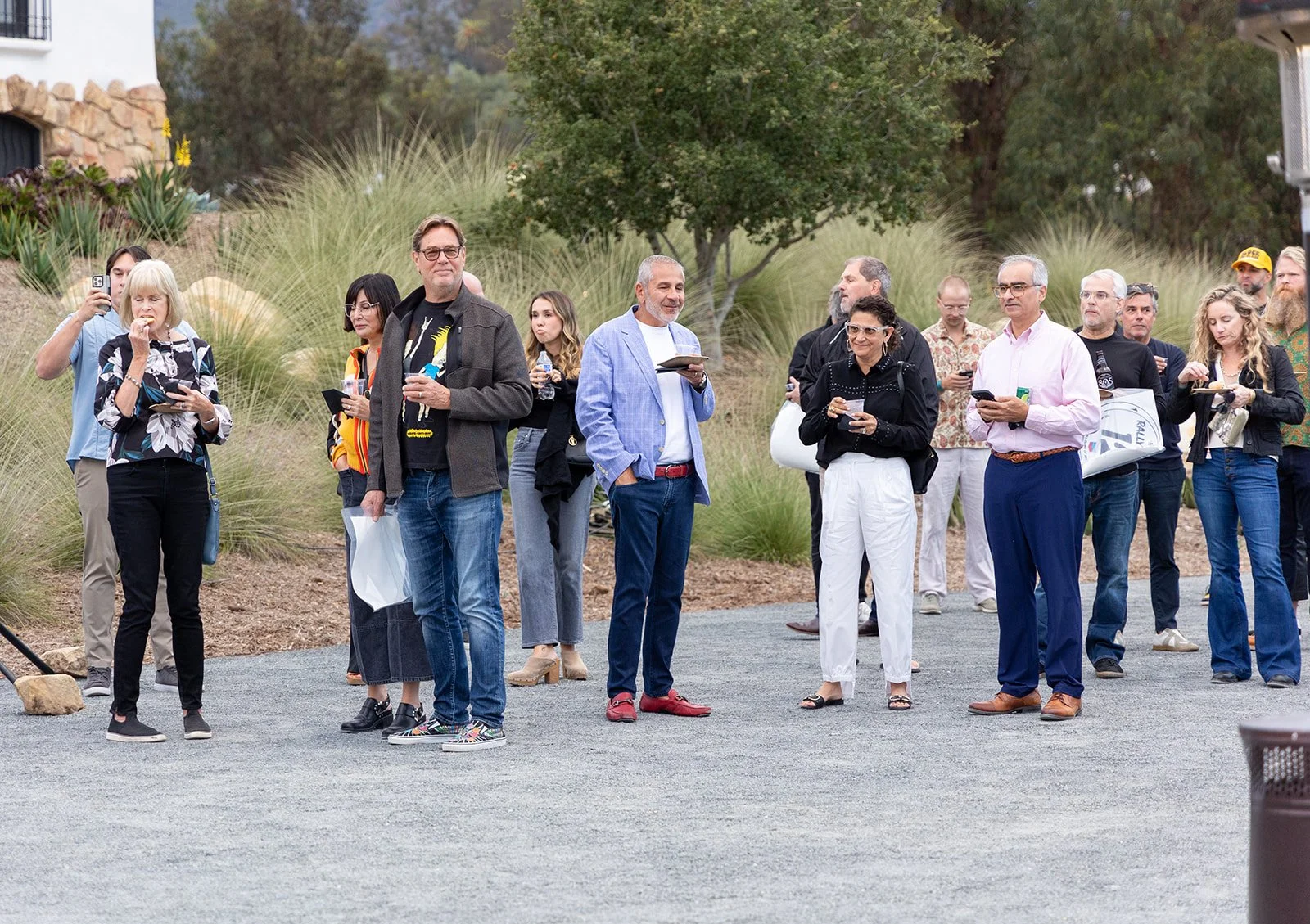 A group of people standing outdoors on a gravel surface, some eating, drinking, or looking at their phones, with grass, trees, and a building in the background.