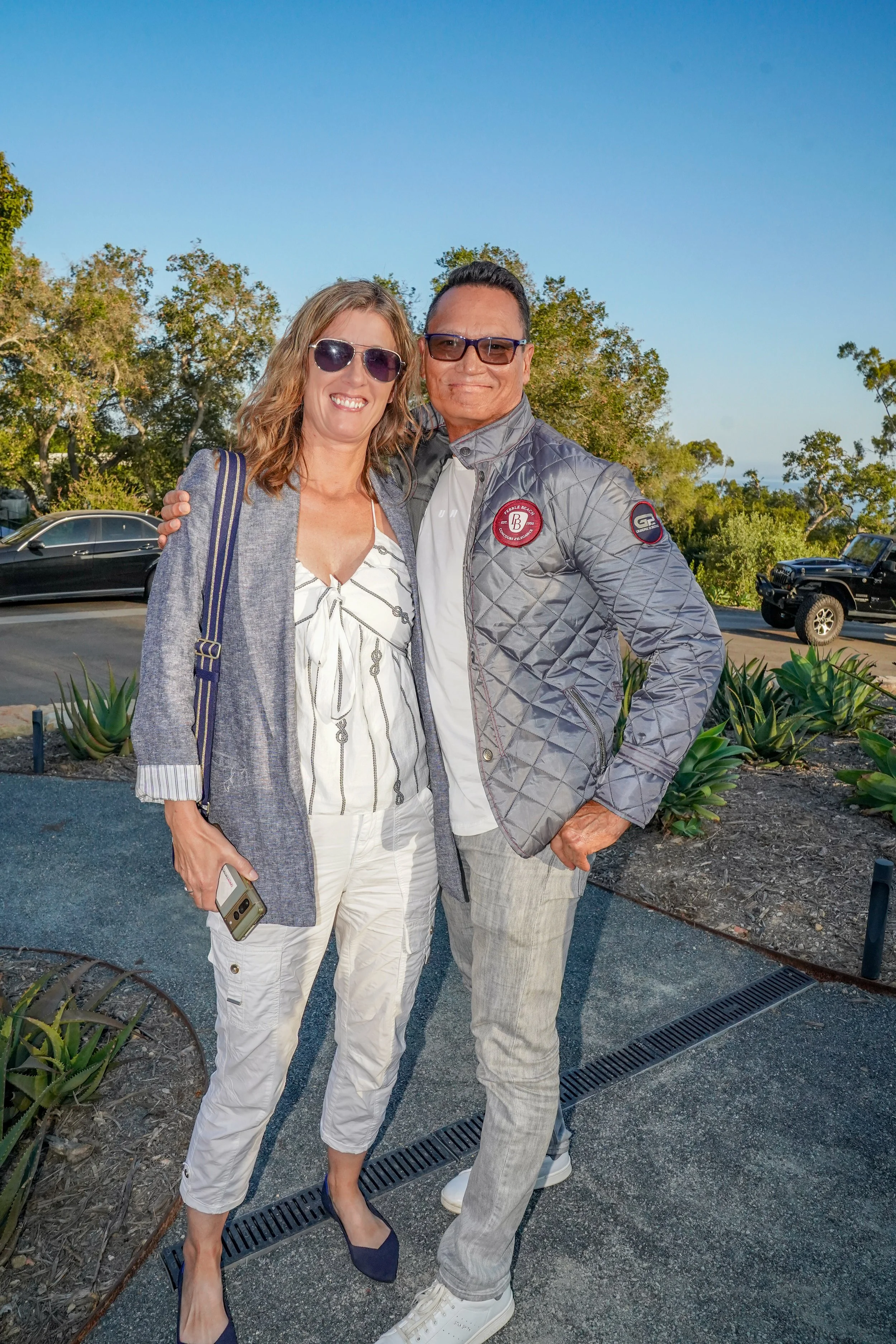 Smiling woman and man standing outdoors on a sunny day, embracing, with greenery and parked cars in the background.
