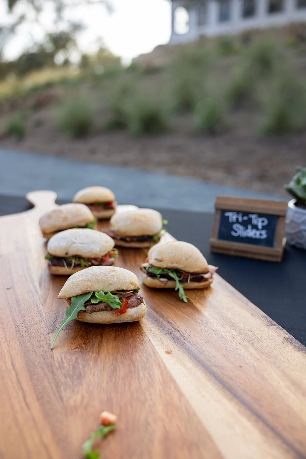 Six small sandwiches with various meats and greens on a wooden serving board outdoors, with a small chalkboard sign reading 'Tri-Tip Sliders' in the background.