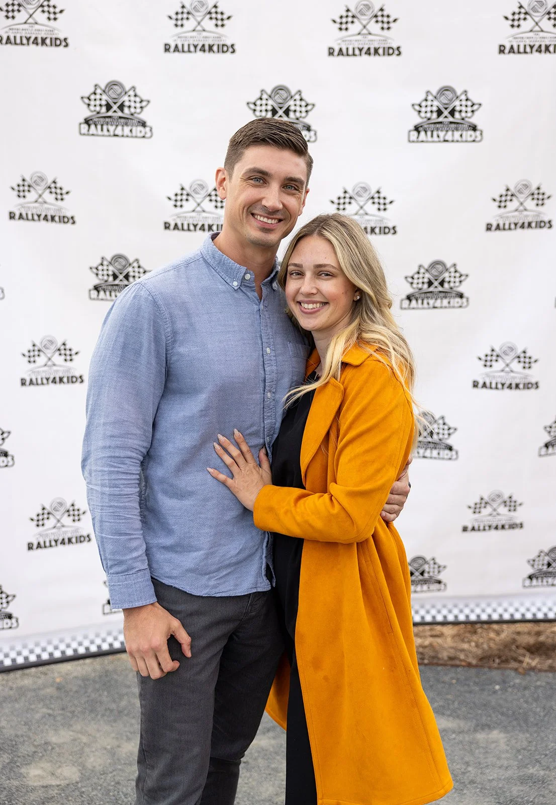 A smiling man and woman standing close together at a rally event, with a backdrop that reads 'Rally4Kids' and features race flags and checkered patterns.