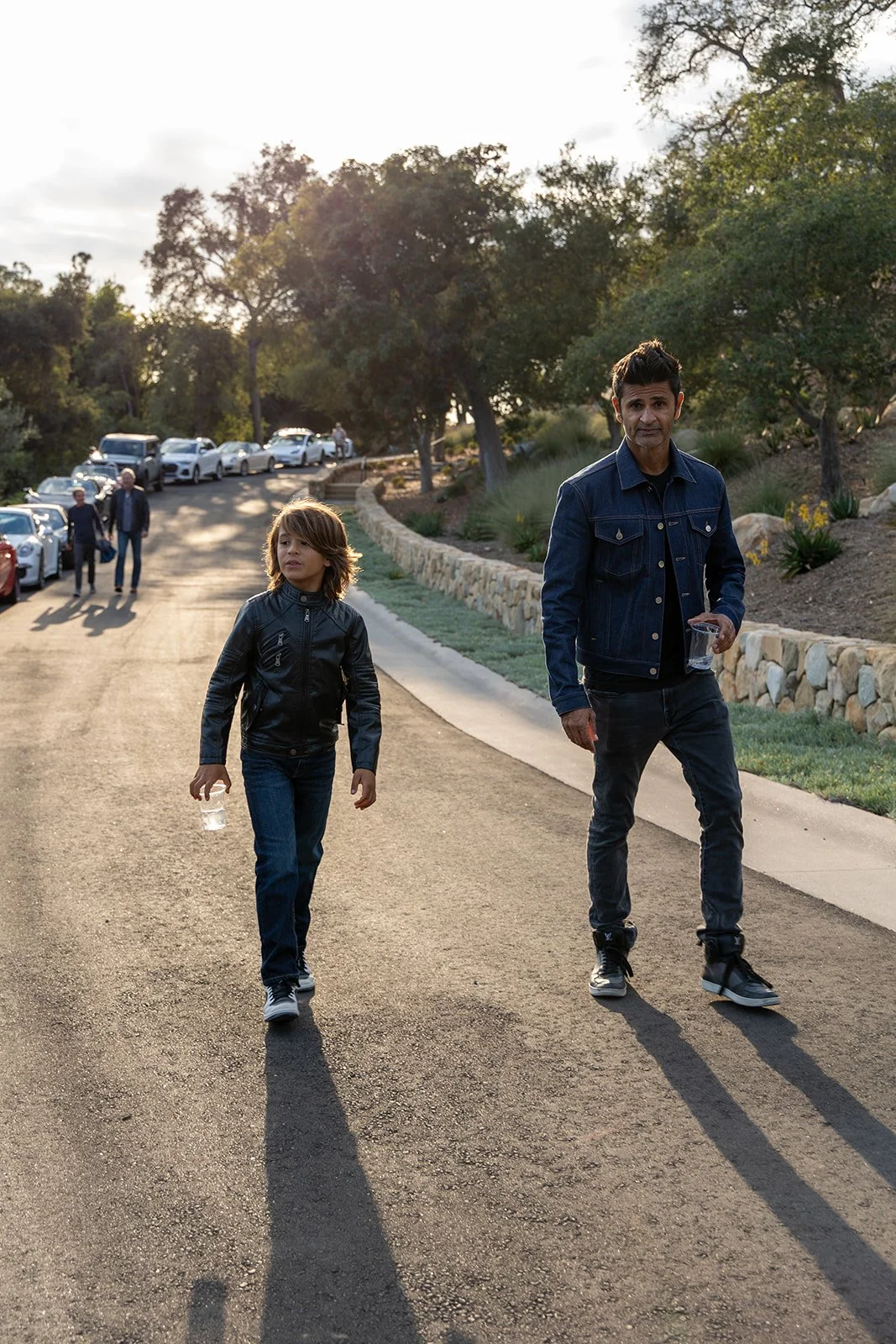 Two young men walking on a paved road during sunset, with parked cars and trees in the background.