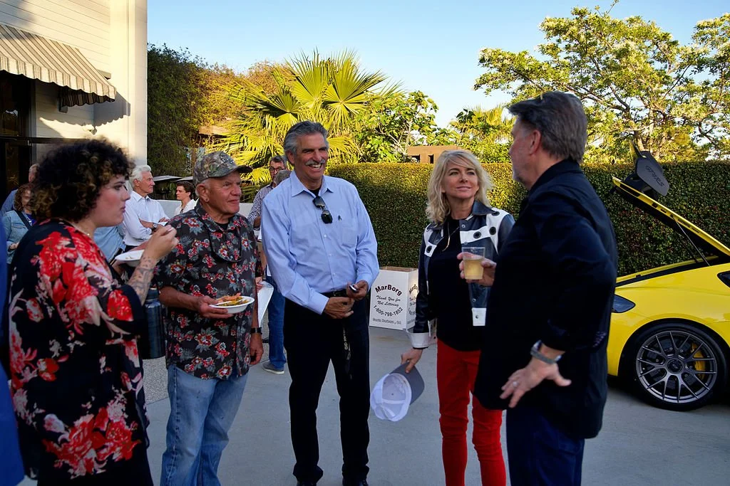 Group of five people casually talking and enjoying drinks outdoors during a social event, with a yellow sports car with its door open in the background.