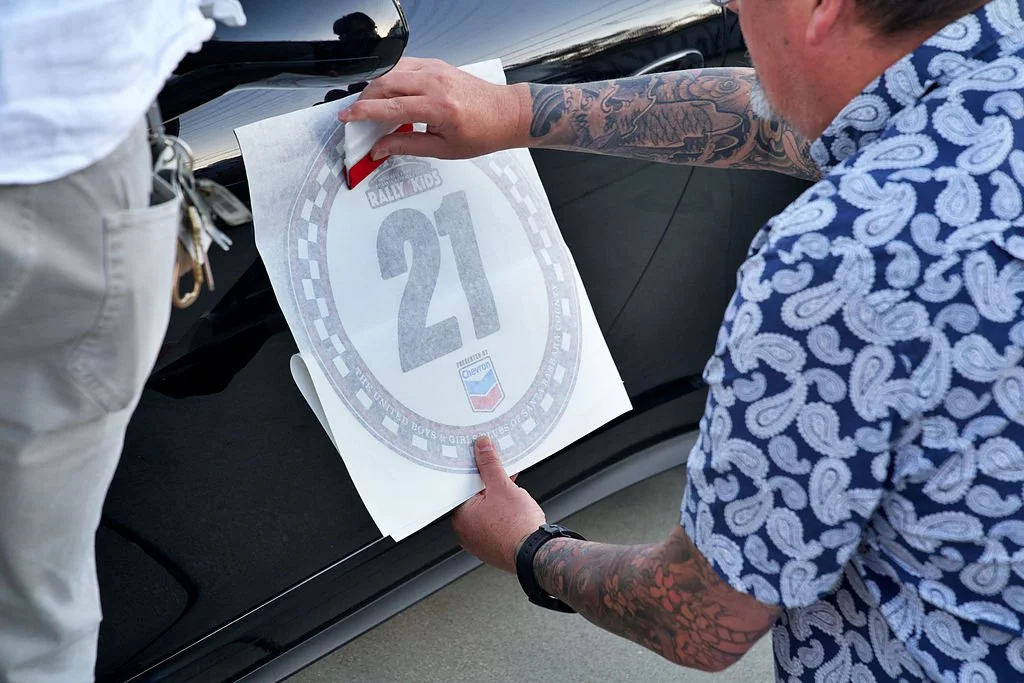 Two people apply a race car decal with the number 21 onto a black car, with one person wearing a blue and white paisley shirt and the other wearing beige pants.