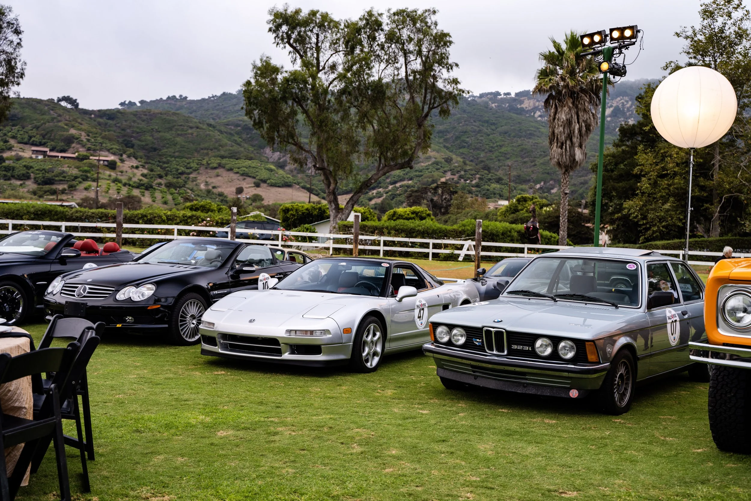 Lineup of classic cars at a car show, including a black Mercedes convertible, a white Chevrolet Corvette, and a gray BMW 3 Series, with a scenic hillside background and decorative lighting.