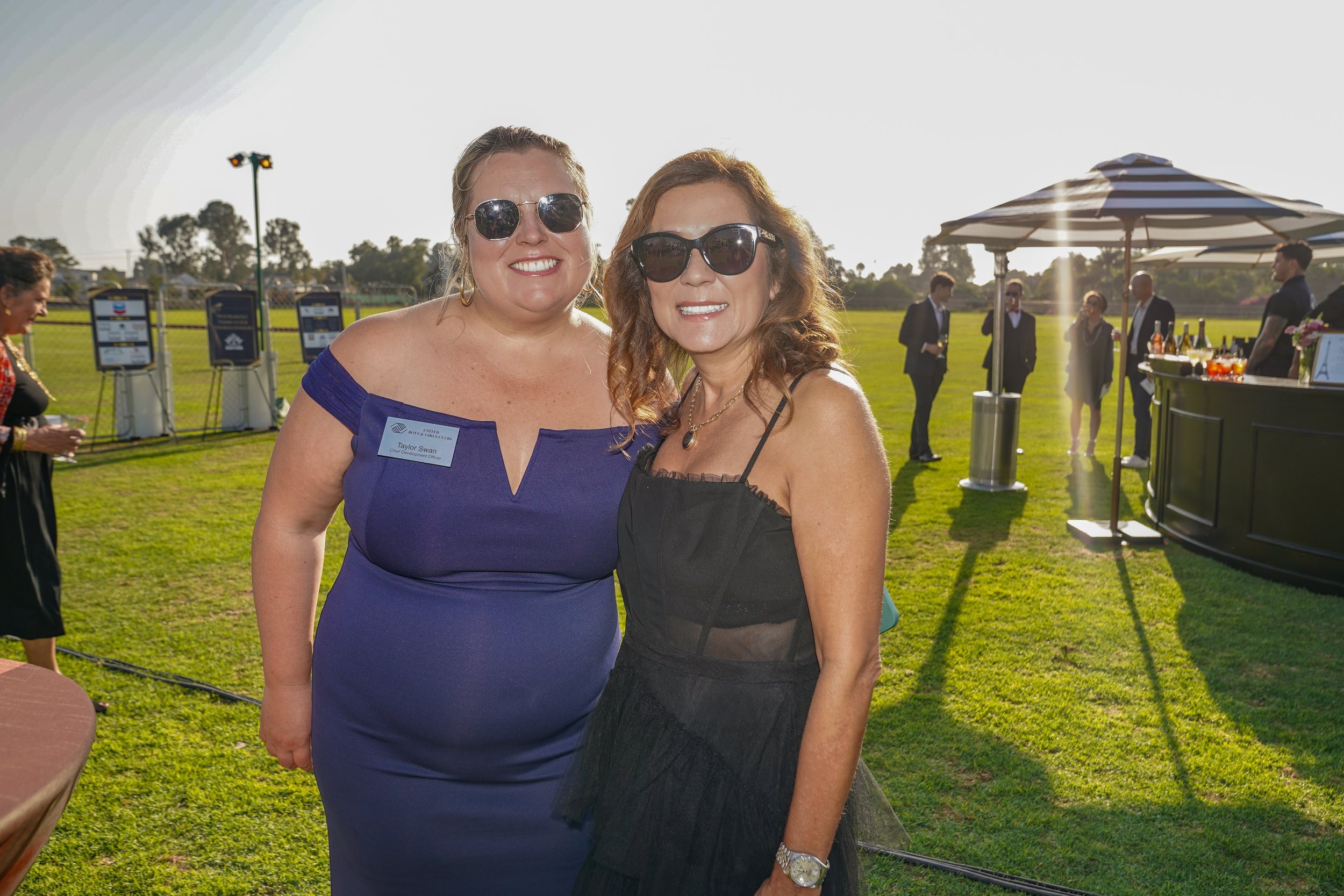 Two women in evening attire wearing sunglasses, standing outdoors on a sunny day, smiling at the camera at a social event on a grassy field with a bar and other guests in the background.