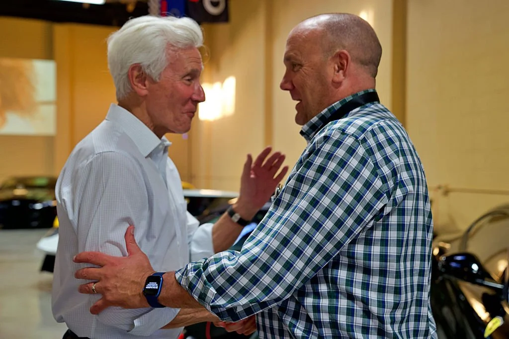 Two men engaging in an emotional conversation indoors, with one touching the other's arm and the other raising his hand slightly.