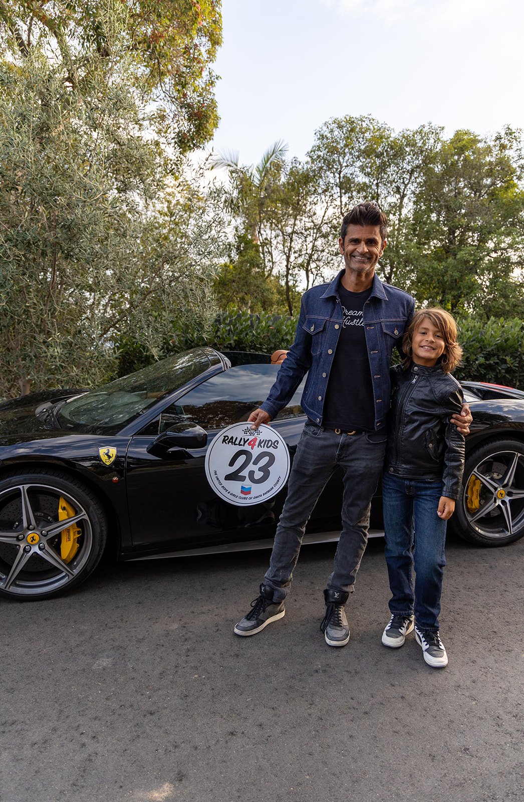 A man and boy smiling and standing in front of a black Ferrari with yellow brake calipers during a rally event. The man is holding a circular sign with the text ‘Rally 4 Kids’ and the number 23.