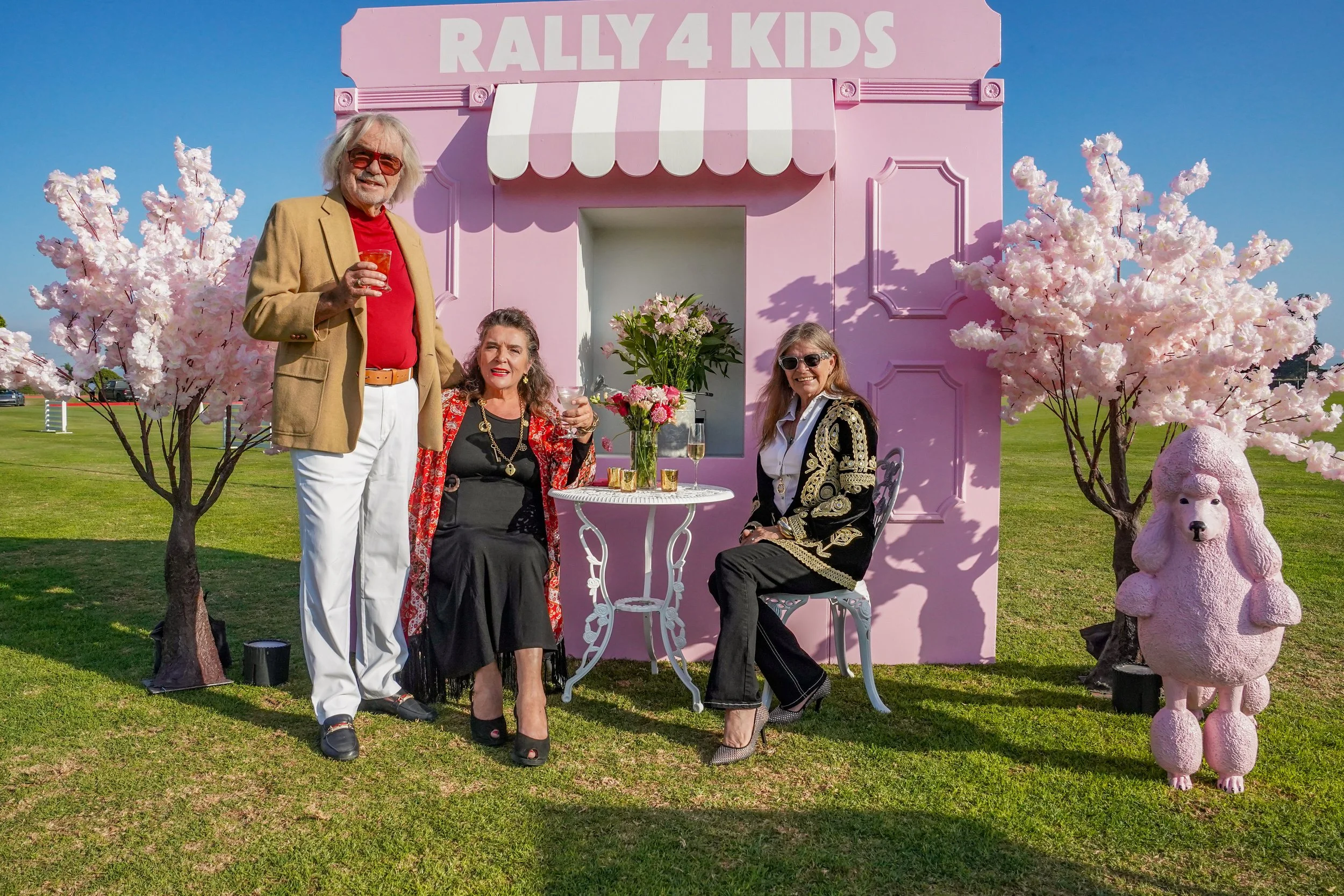 Three people celebrating outdoors in front of a pink building labeled 'Rally 4 Kids', with pink cherry blossom trees and a pink poodle sculpture. They are holding drinks, with a table of flowers and champagne in front of them.