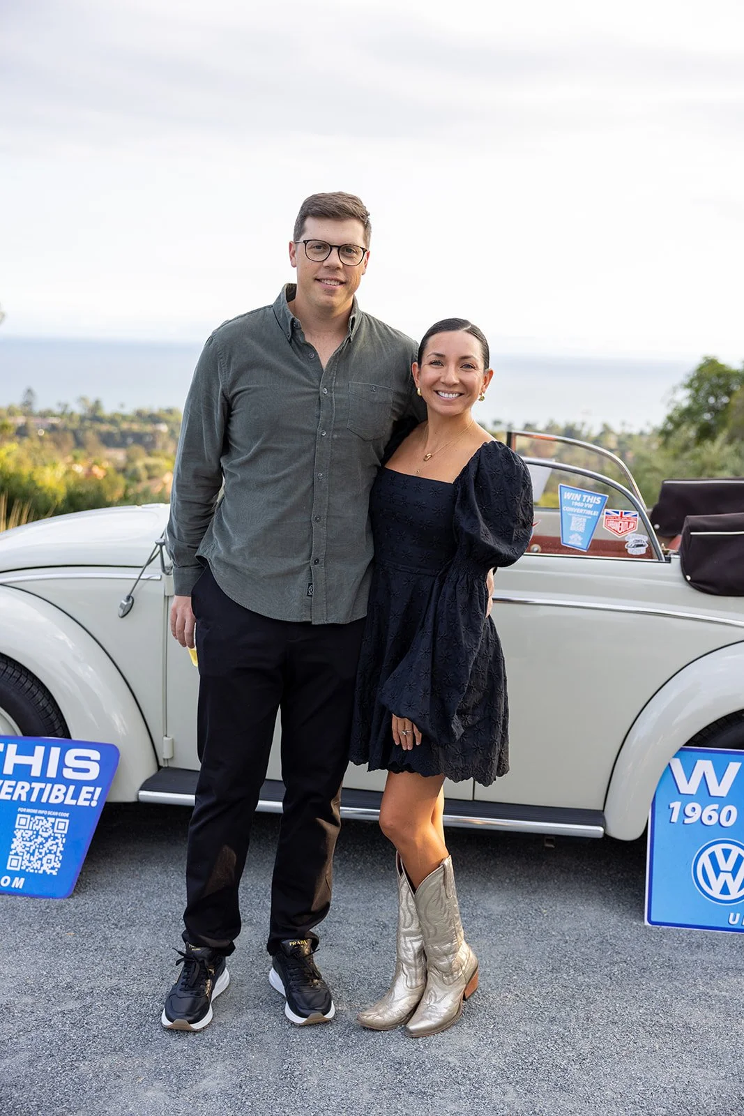 A man and woman standing together in front of a vintage VW Beetle, smiling at the camera, with outdoor scenery and promotional signs nearby.