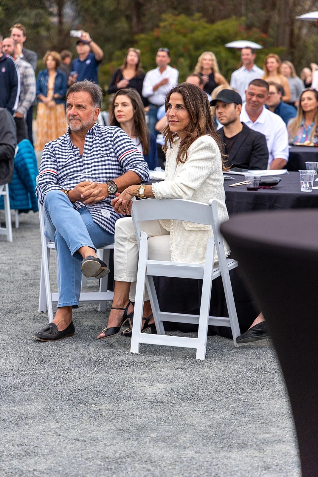 A group of people attending an outdoor event, seated at tables with some standing in the background, with trees and foliage behind them.