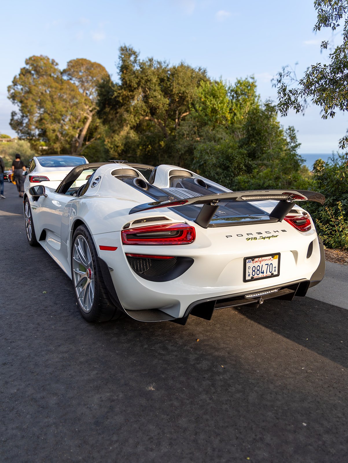 A white Porsche 918 Spyder with a large rear wing parked on the side of a road, with trees and a blue sky in the background.
