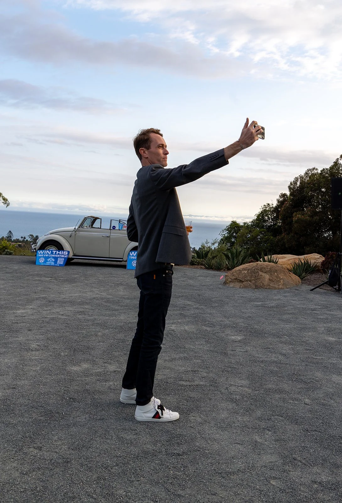 A man in a dark blazer and black jeans taking a selfie on a gravel surface near a vintage Volkswagen convertible car with a sign that says "Win This". There are trees, rocks, and an ocean view in the background during sunset or late afternoon.