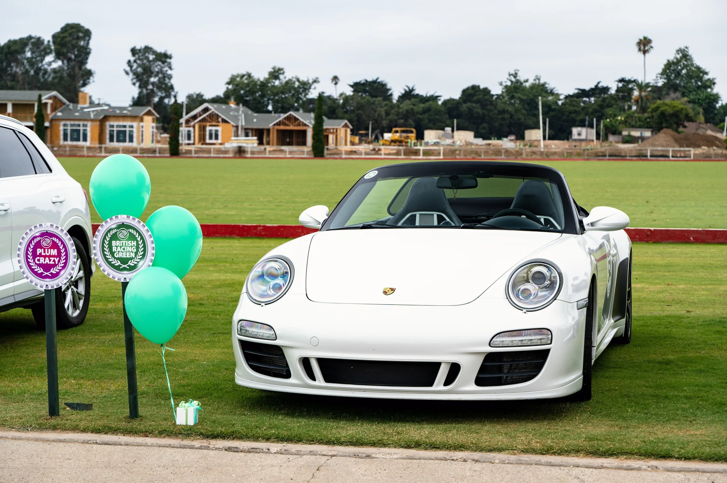A white Porsche convertible parked on a grassy field at a car show, with colorful balloons and signs reading 'Plum Crazy' and 'British Racing Green' next to it.