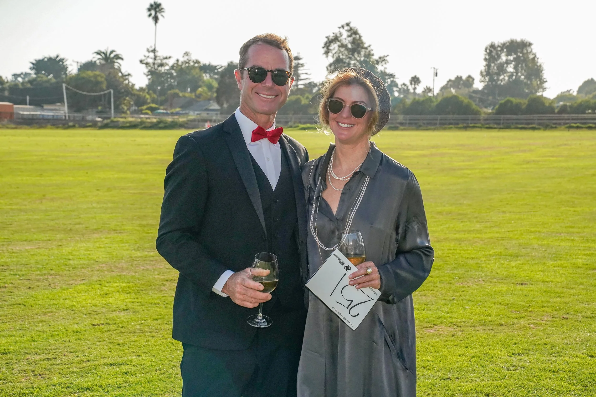 A man and woman dressed in formal attire standing outdoors on a grassy field, holding wine glasses, smiling at the camera with trees and a clear sky in the background.