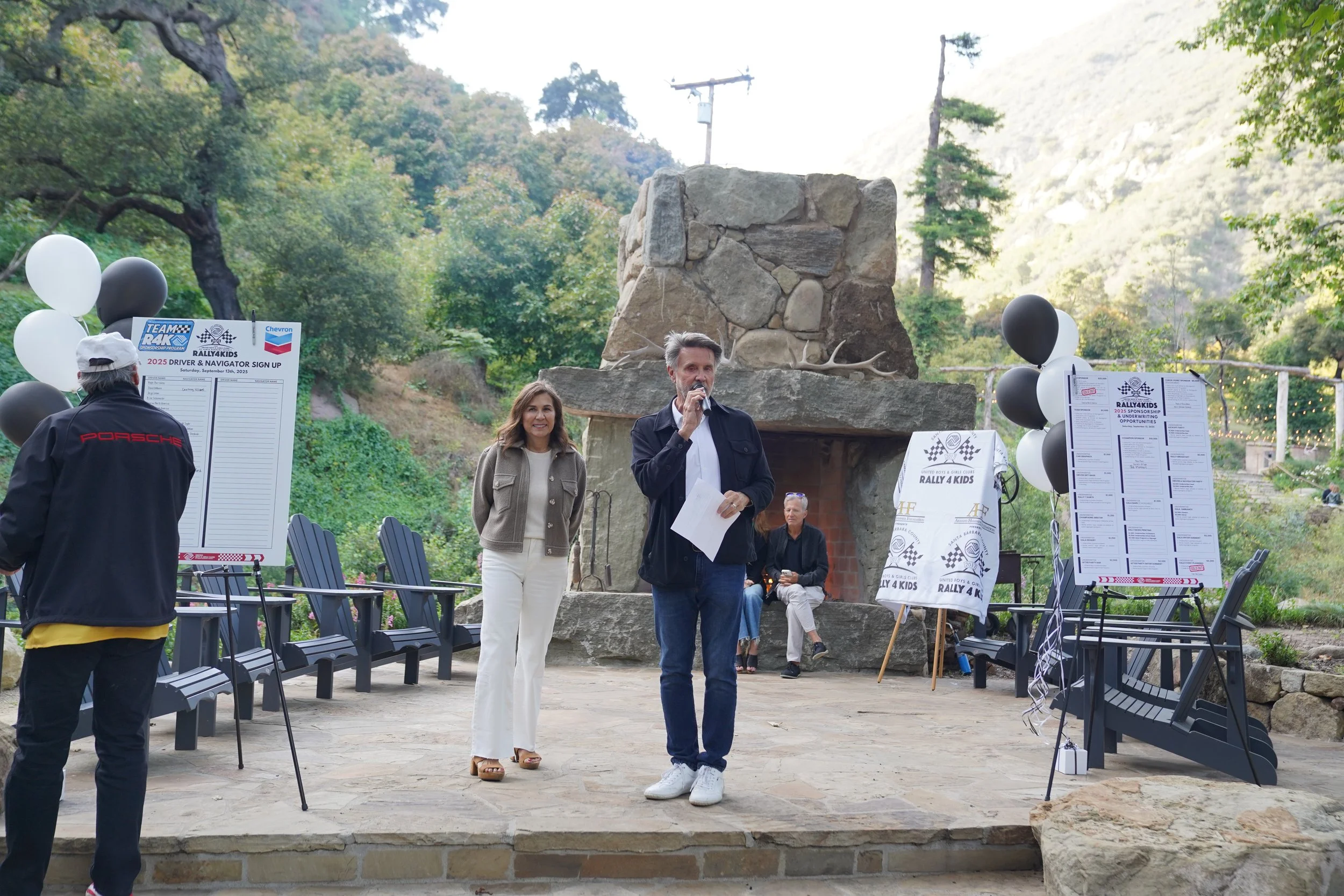 People gathered outdoors at a rally event with signs, balloons, and a fireplace backdrop surrounded by trees and mountains.