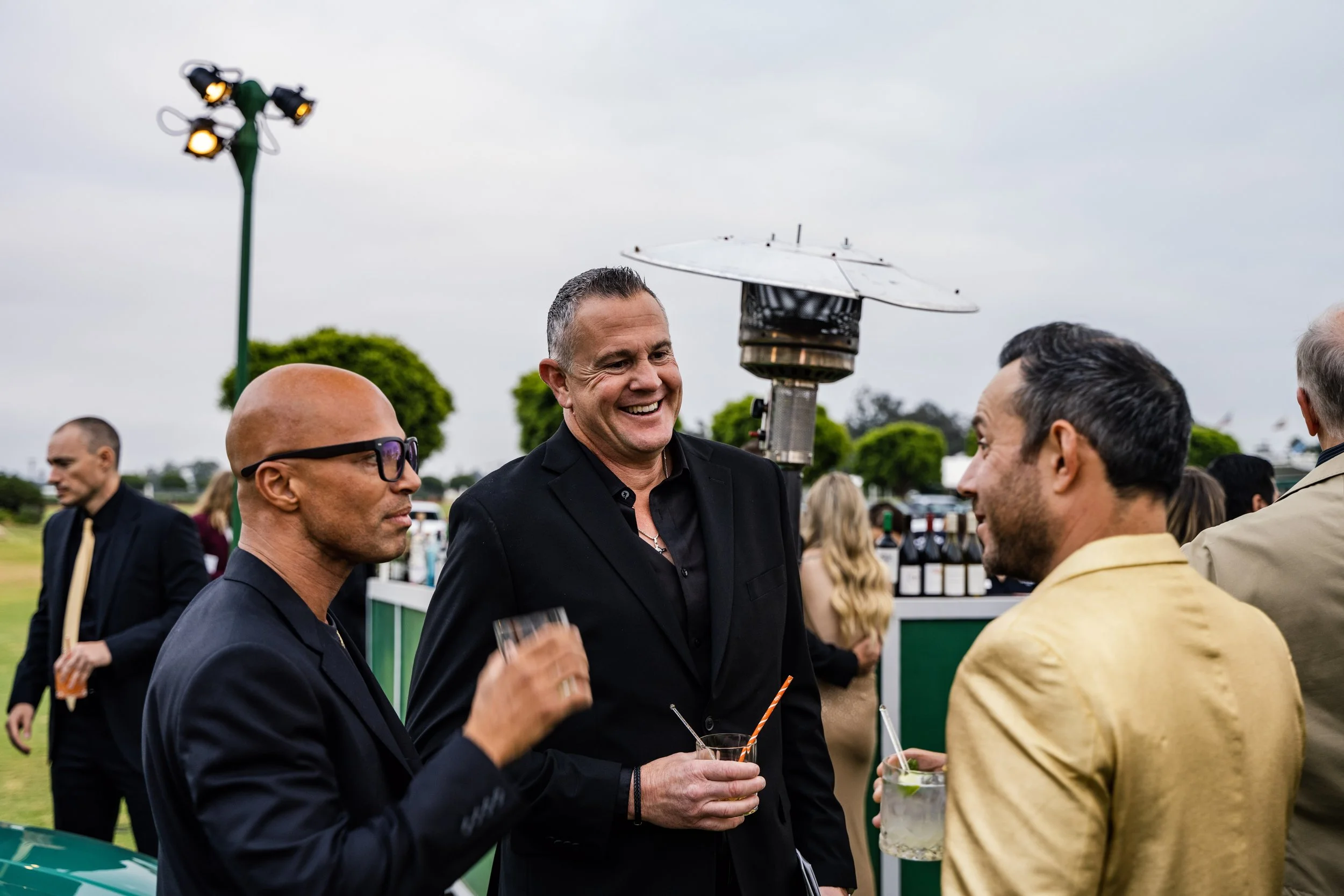 People socializing outdoors at a gathering, with a man in a black suit smiling, holding a drink, and a man in a tan jacket talking, with others in the background and outdoor lighting.