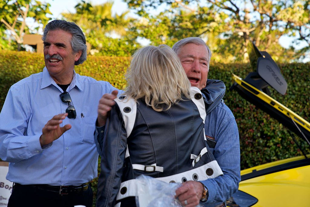 Two men smiling and one emotional person hugging a woman with a backpack outdoors with a yellow sports car in the background.