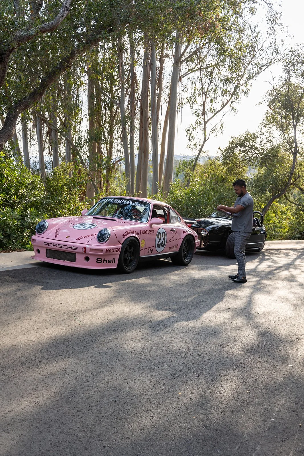 A pink Porsche racing car with number 23 and various sponsor logos parked on a road, with a man standing beside it near a black car. The background features trees and sunlight filtering through the leaves.