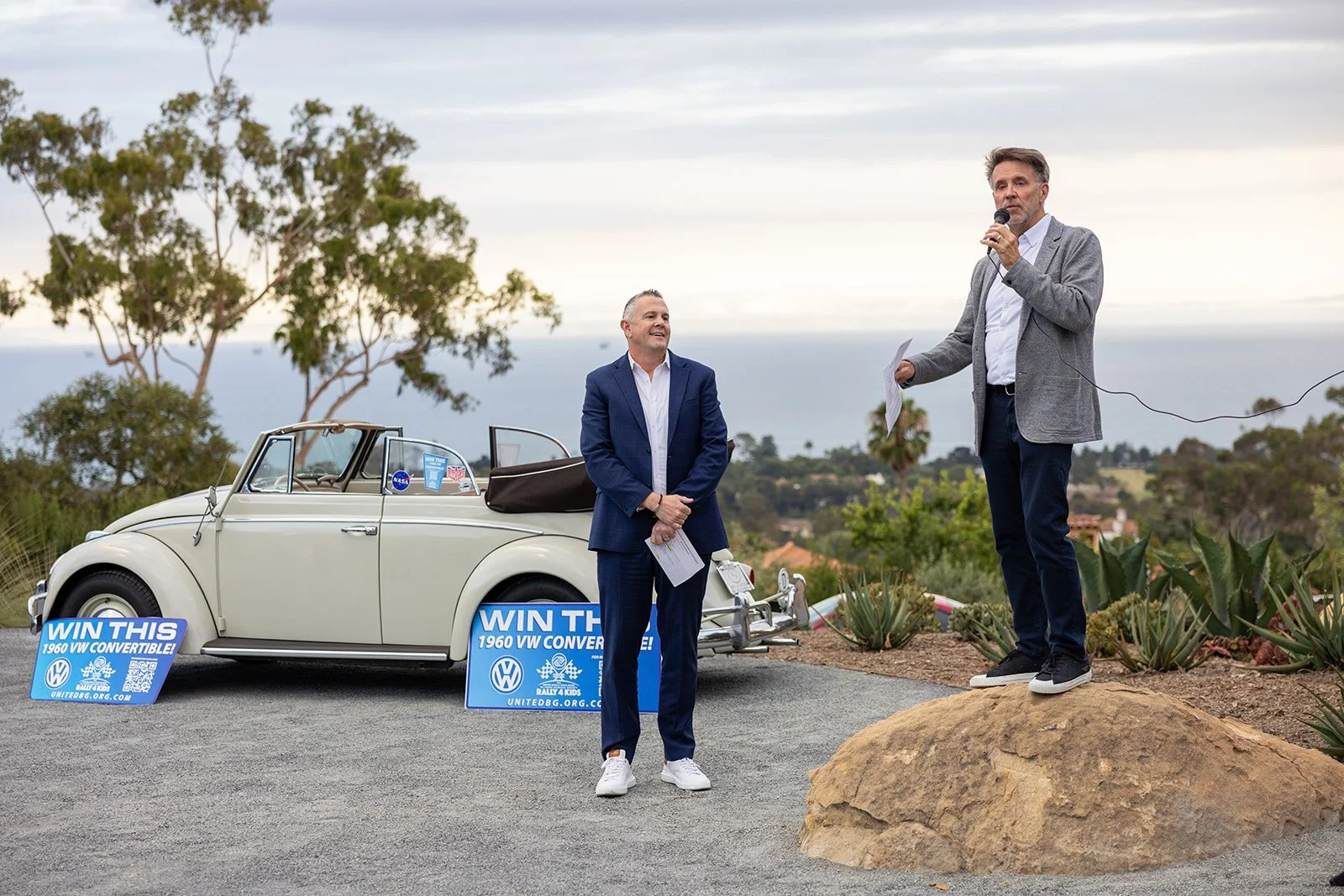 Two men are outdoors during daytime, with one man in a gray blazer standing on a rock and speaking into a microphone, and another man in a blue suit standing nearby holding papers. There is a vintage white convertible car with signs in front of it th