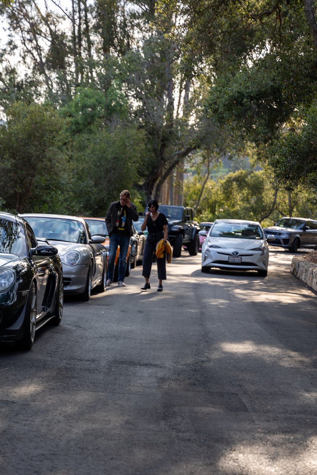 Two people walking along a parking lot filled with parked cars, surrounded by trees with sunlight filtering through.