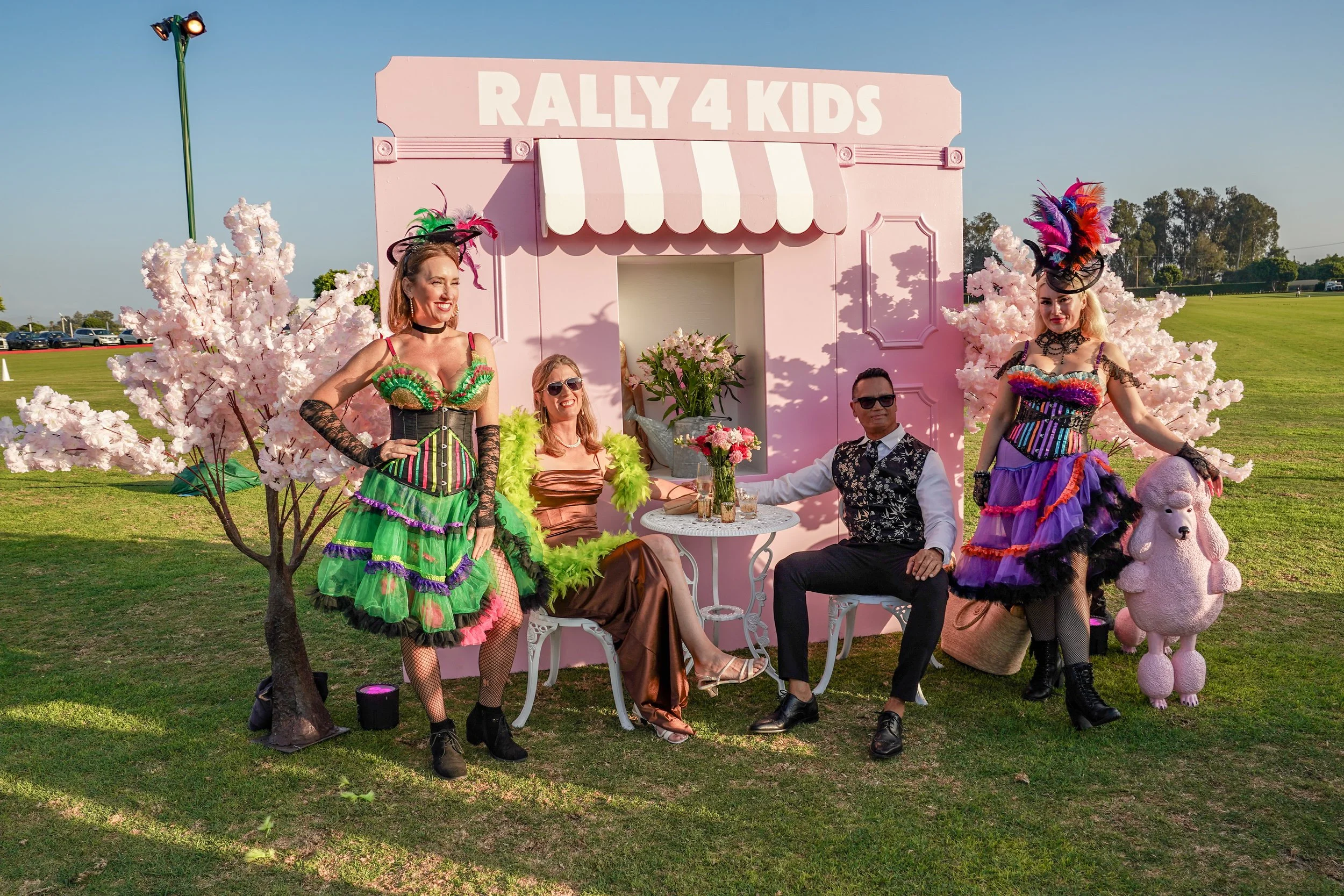 Four people dressed in colorful, theatrical costumes sit at a small, round white table outside, surrounded by pink cherry blossom trees. Behind them is a pink wall with a sign that says "RALLY 4 KIDS". Two of the women are standing, wearing vibrant d