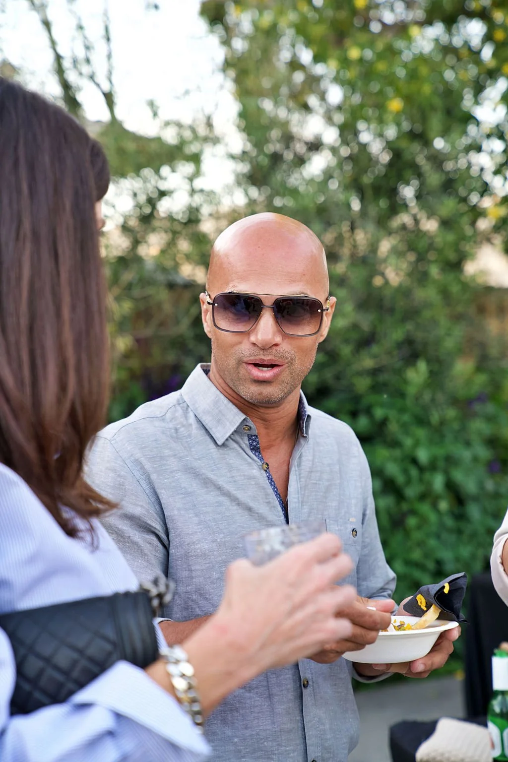 A bald man wearing sunglasses and a light gray shirt is holding a bowl of food, talking to a woman in a blue shirt with a black quilted jacket on her arm. They are outdoors with green trees in the background.