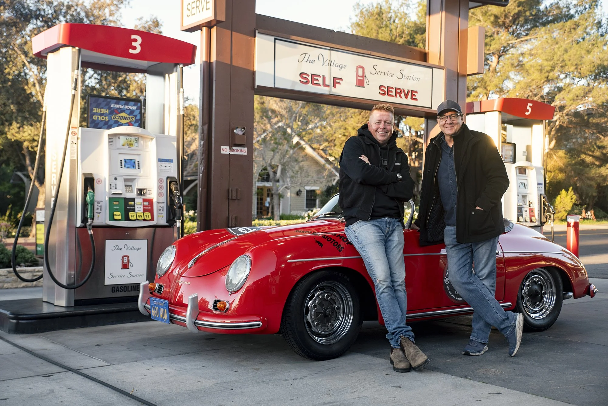 Two men in black jackets and jeans are standing beside a red vintage sports car parked at a gas station with self-serve pumps. They are smiling and leaning against the car, which is under a sign that reads 'The Village Service Station'.