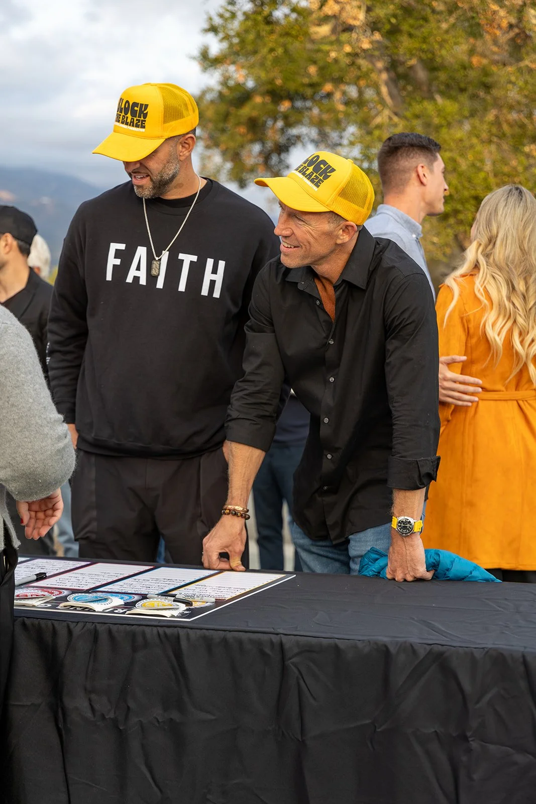 Two men wearing yellow hats with black text, engaging around a table outdoors during daytime, with other people and autumn trees in the background.