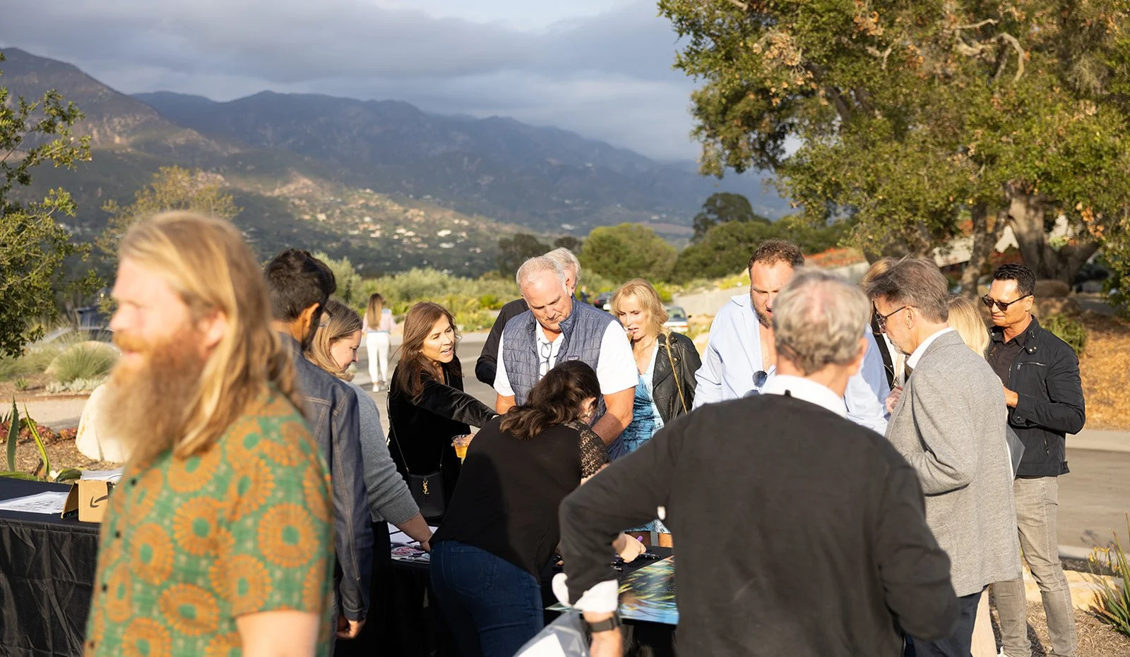 A group of people gathered outdoors near a table, with mountains and trees in the background.