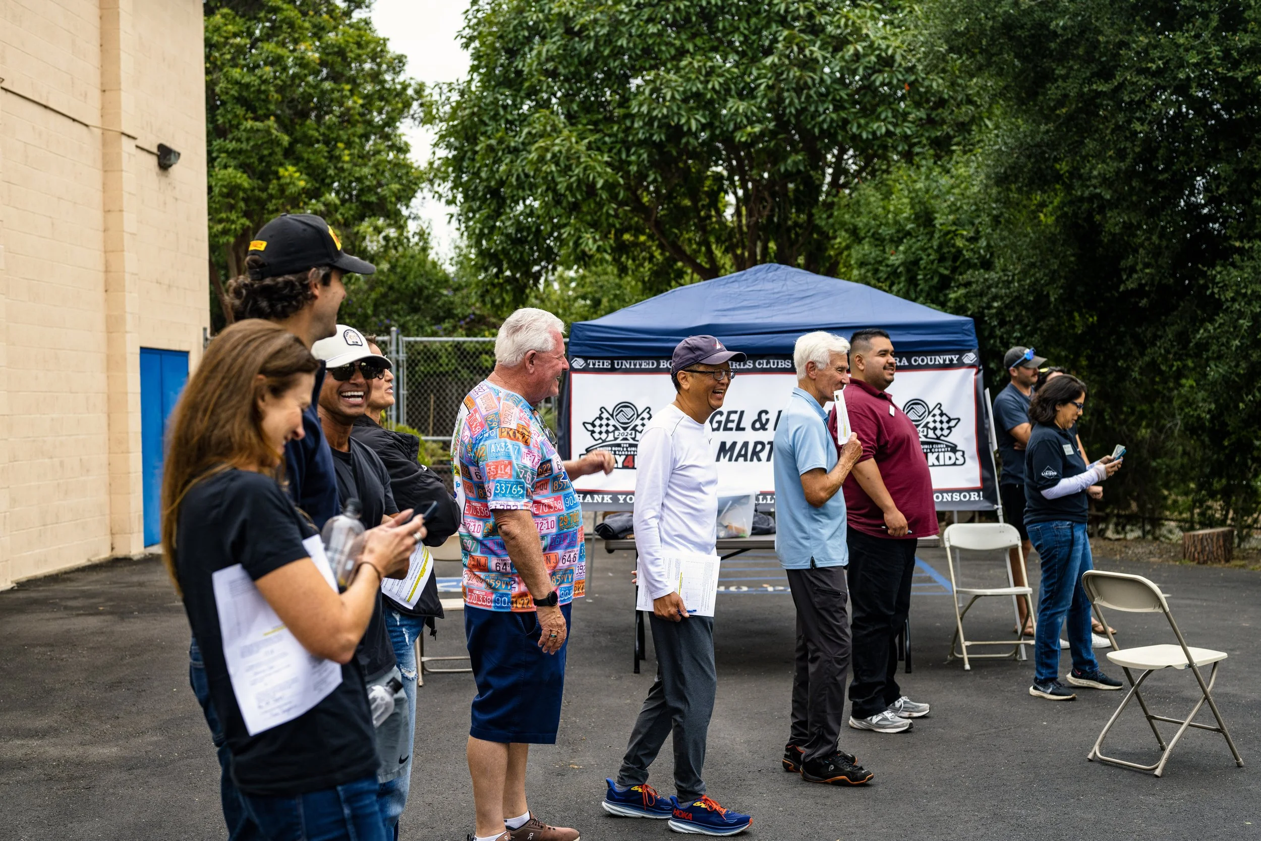 Group of people standing in line outdoors near a blue tent, some holding papers or mobile phones, participating in a community event with trees in the background.