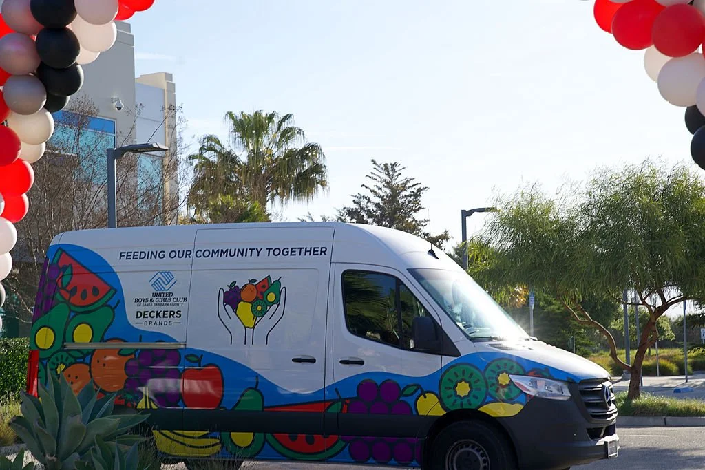 A white van decorated with colorful fruit illustrations and the logo of the Boys & Girls Clubs of San Gabriel Valley, parked under a balloon arch with red, white, and black balloons. The van has the phrase "Feeding Our Community Together" written on 