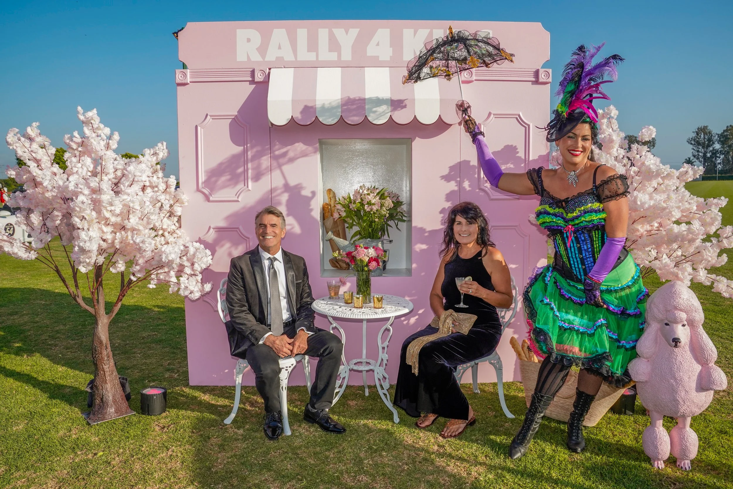 Group of three people sitting and standing in front of a pink building with a sign that says 'Rally for Kh' on it, surrounded by pink cherry blossom trees and decorative pink poodle statues. One person dressed in colorful costume stands to the right,