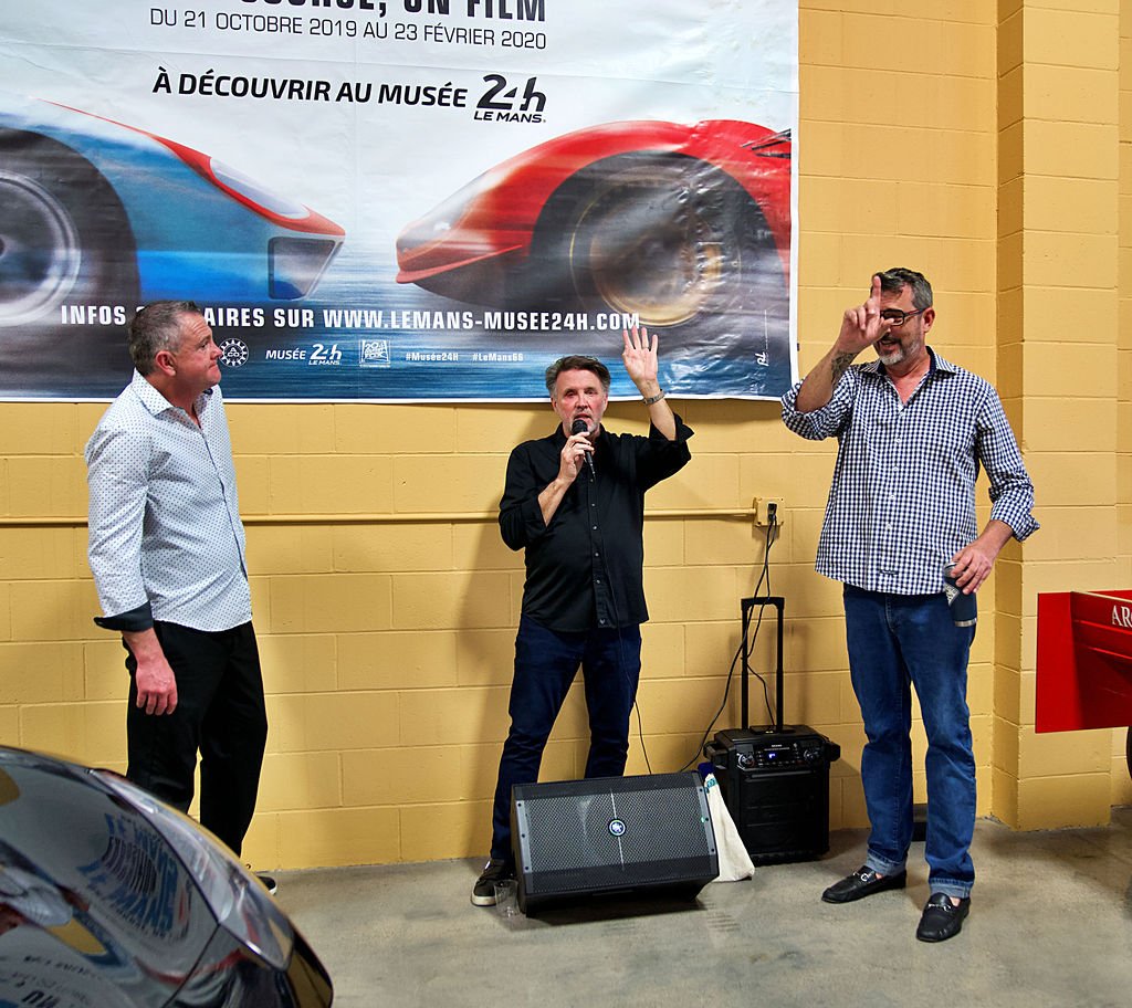 Three men standing and talking at an indoor event in front of a large advertisement banner for a car exhibition at a museum. One man is speaking into a microphone, the other two are listening and gesturing.