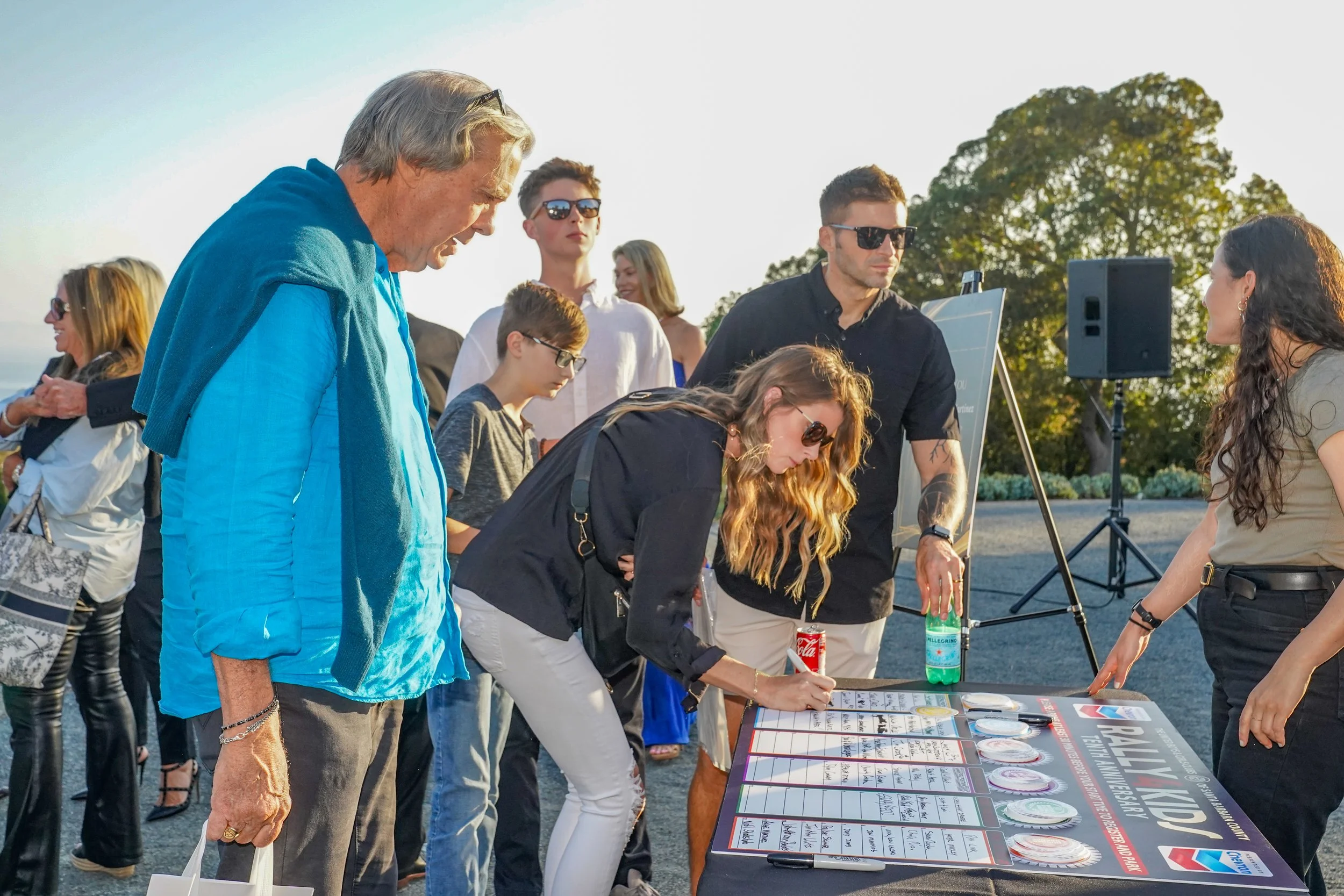 People gathered outdoors at a table for a voting or registration event, with a woman signing a document, various beverages on the table, and others standing in line or observing.