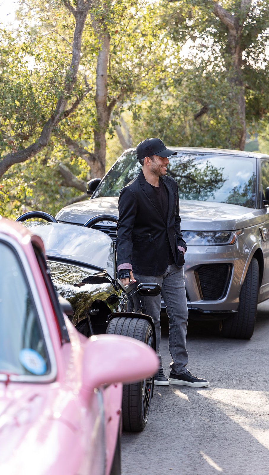 A smiling young man in a black jacket, gray jeans, and a baseball cap standing beside a black sports car, with a silver SUV and trees in the background.