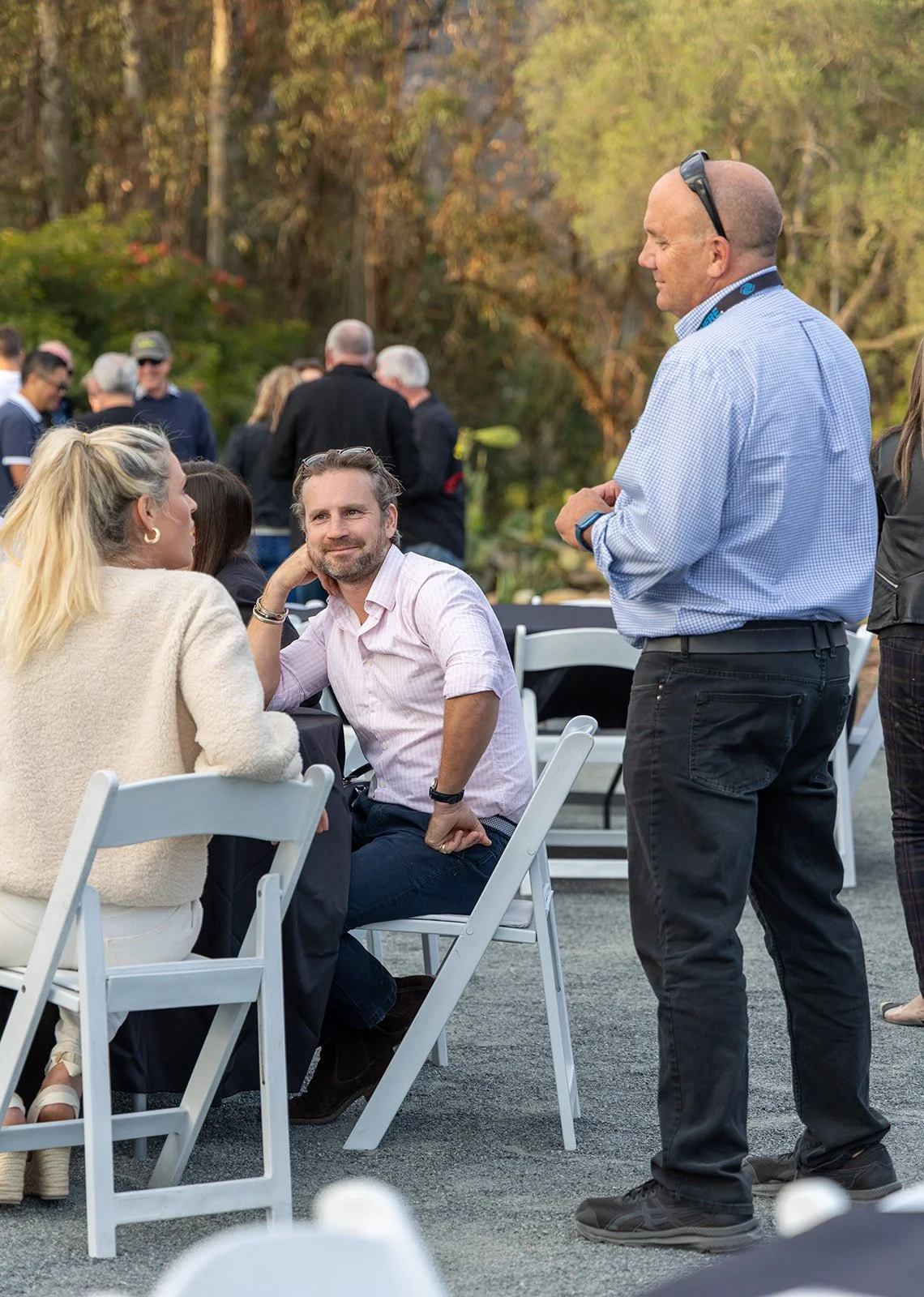 People socializing outdoors at a gathering or event, seated and standing, with trees and foliage in the background.