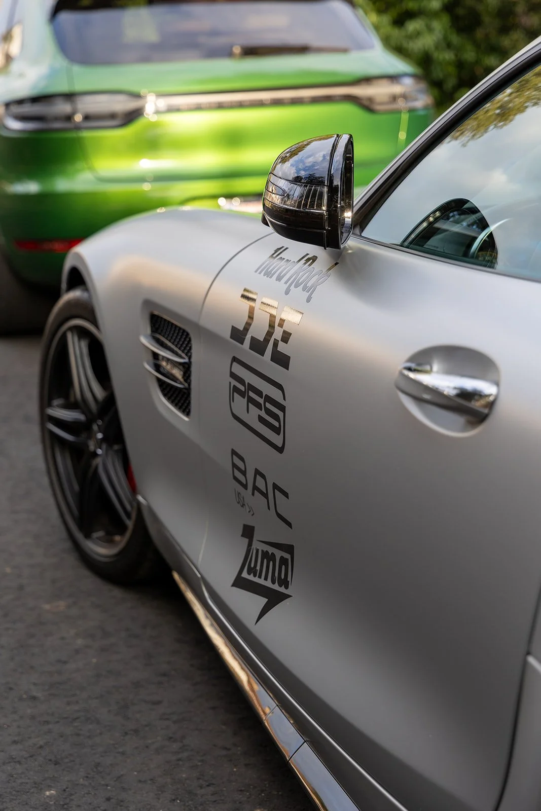 Close-up of a luxury sports car with racing decals, parked next to a green vehicle, on a paved road.