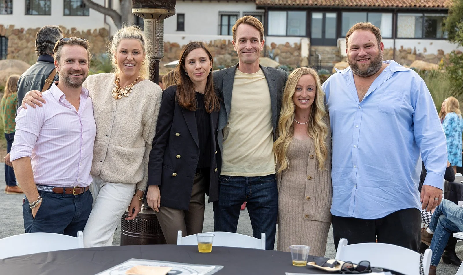 Group of six people standing outdoors, smiling, at a social gathering or party, with a house and other guests in the background.