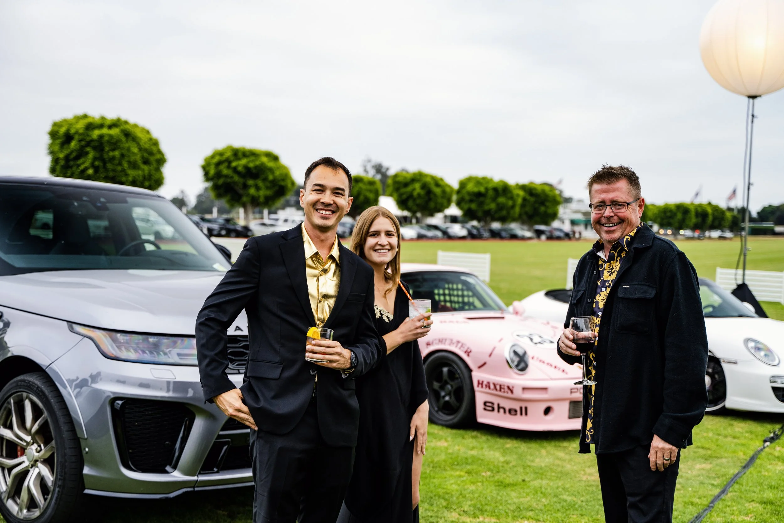 Three people standing outdoors at a car event, smiling and holding drinks, with a silver SUV and a pink race car in the background.