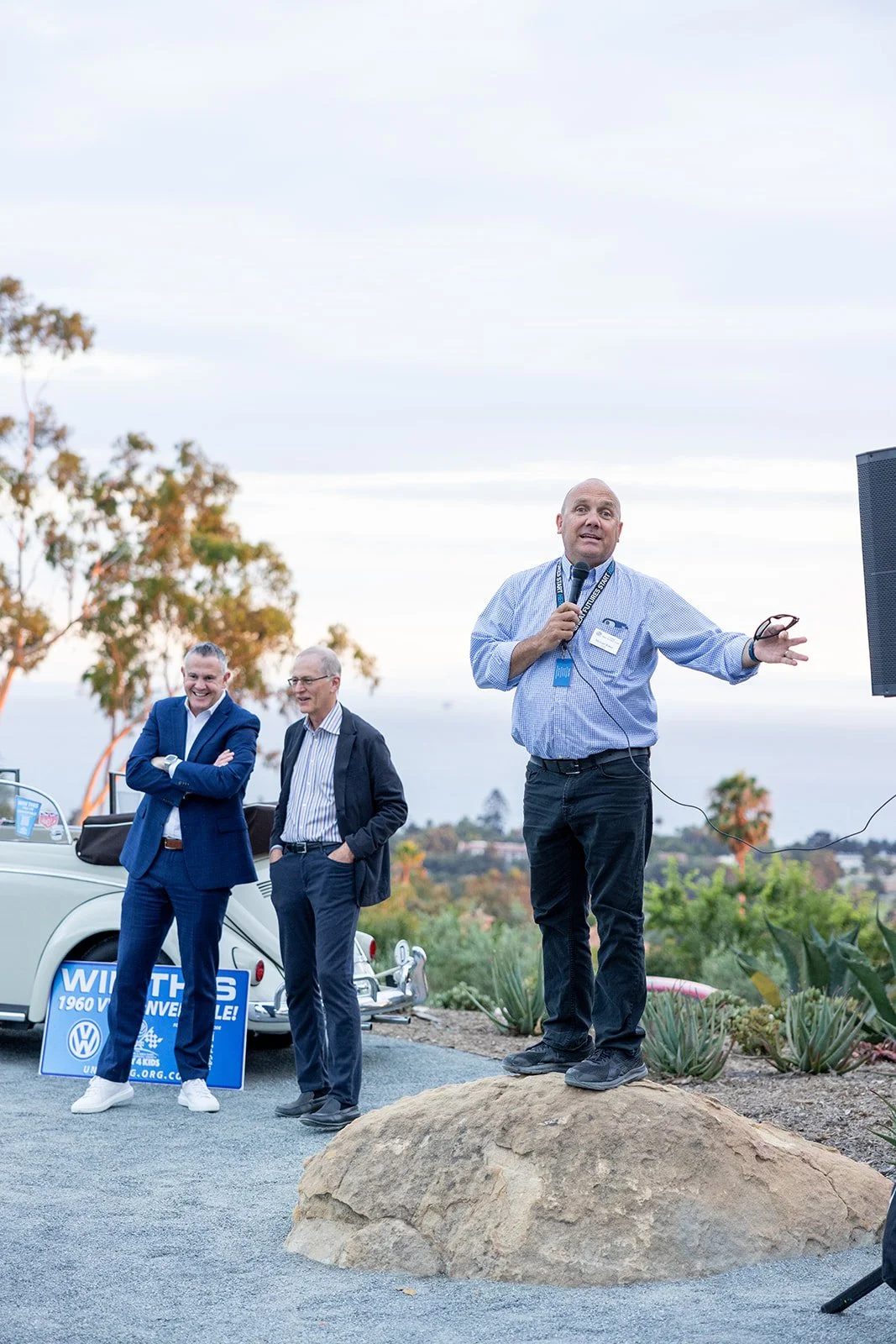 A man speaking into a microphone while standing on a large rock, with two men smiling and standing nearby, outdoors during daytime with desert plants and trees in the background.