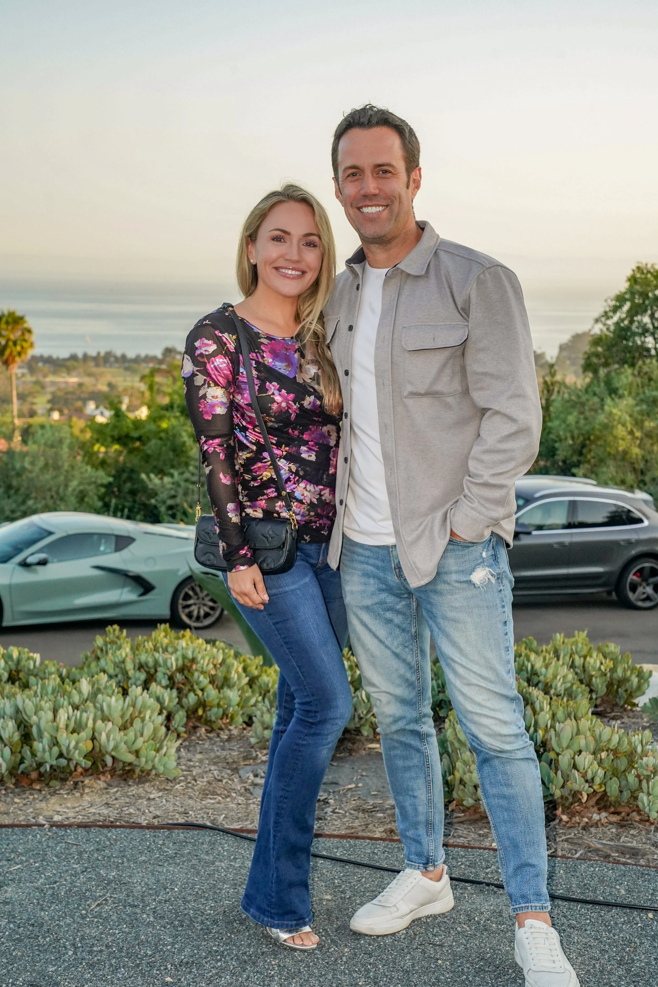 A smiling couple stands outdoors with cars and greenery in the background, during sunset.