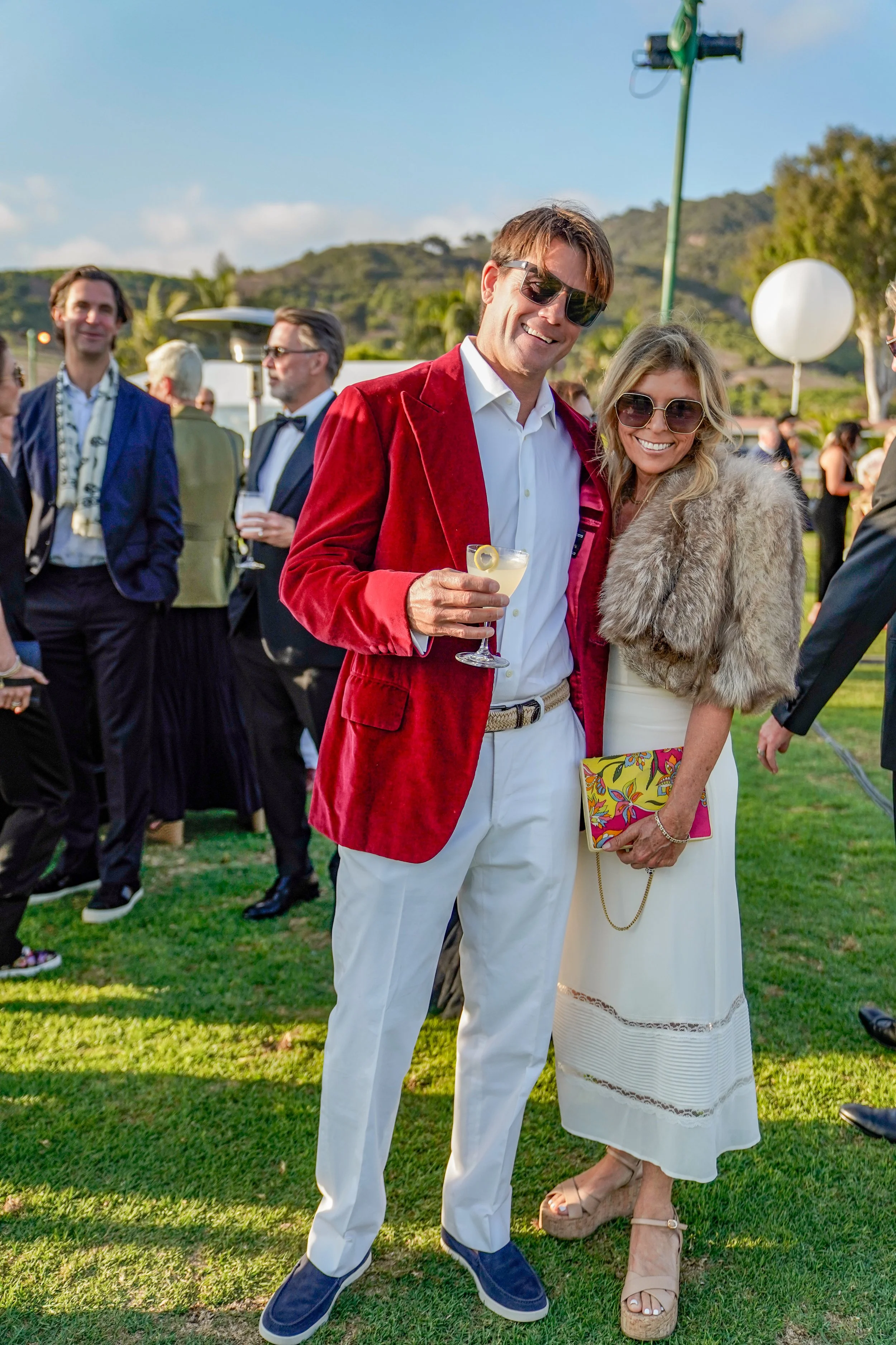 People dressed in formal and stylish clothing at an outdoor event or party, with a man wearing a red velvet blazer and sunglasses, and a woman in a white dress with a fur stole and sunglasses, holding a colorful clutch, standing on a grassy area with