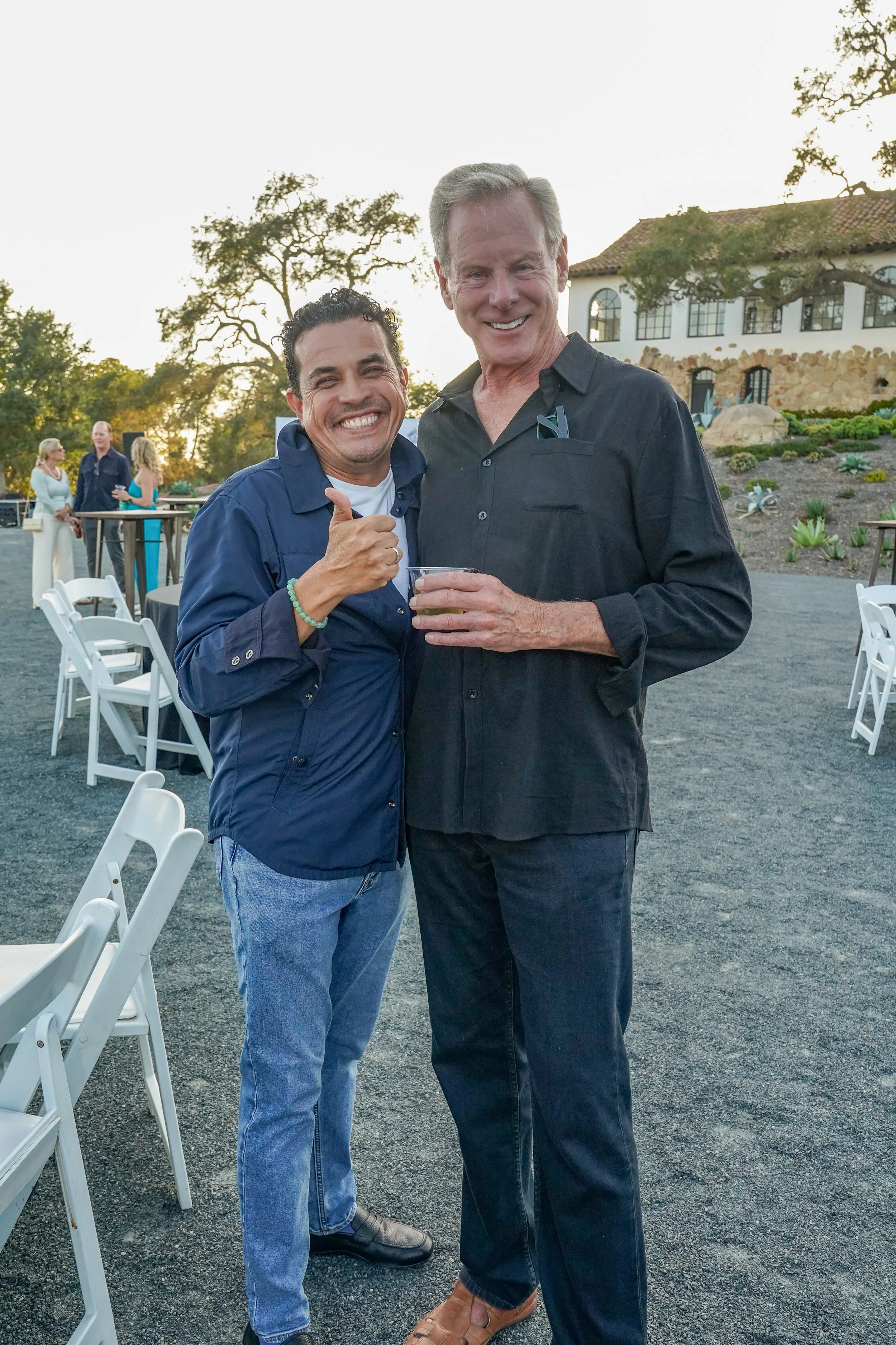 Two men smiling and posing for a photo at an outdoor event during sunset, with white chairs, a building, and other people in the background.