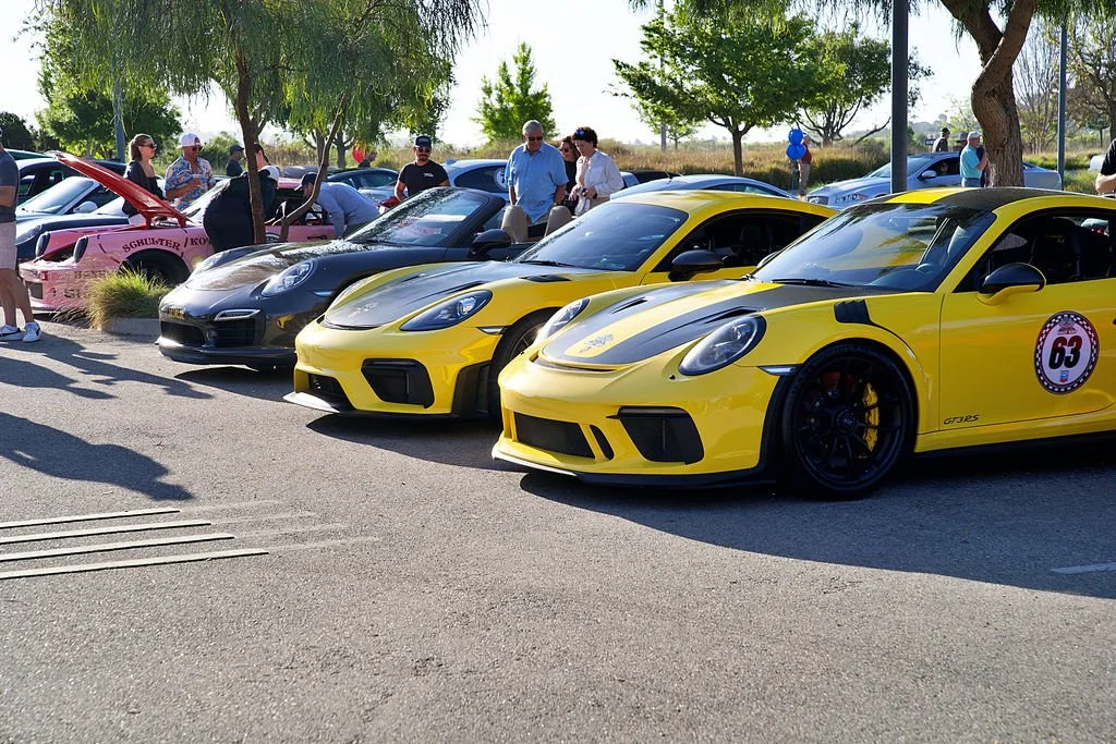 Lineup of yellow and gray sports cars at a car show with people walking around and trees in the background.